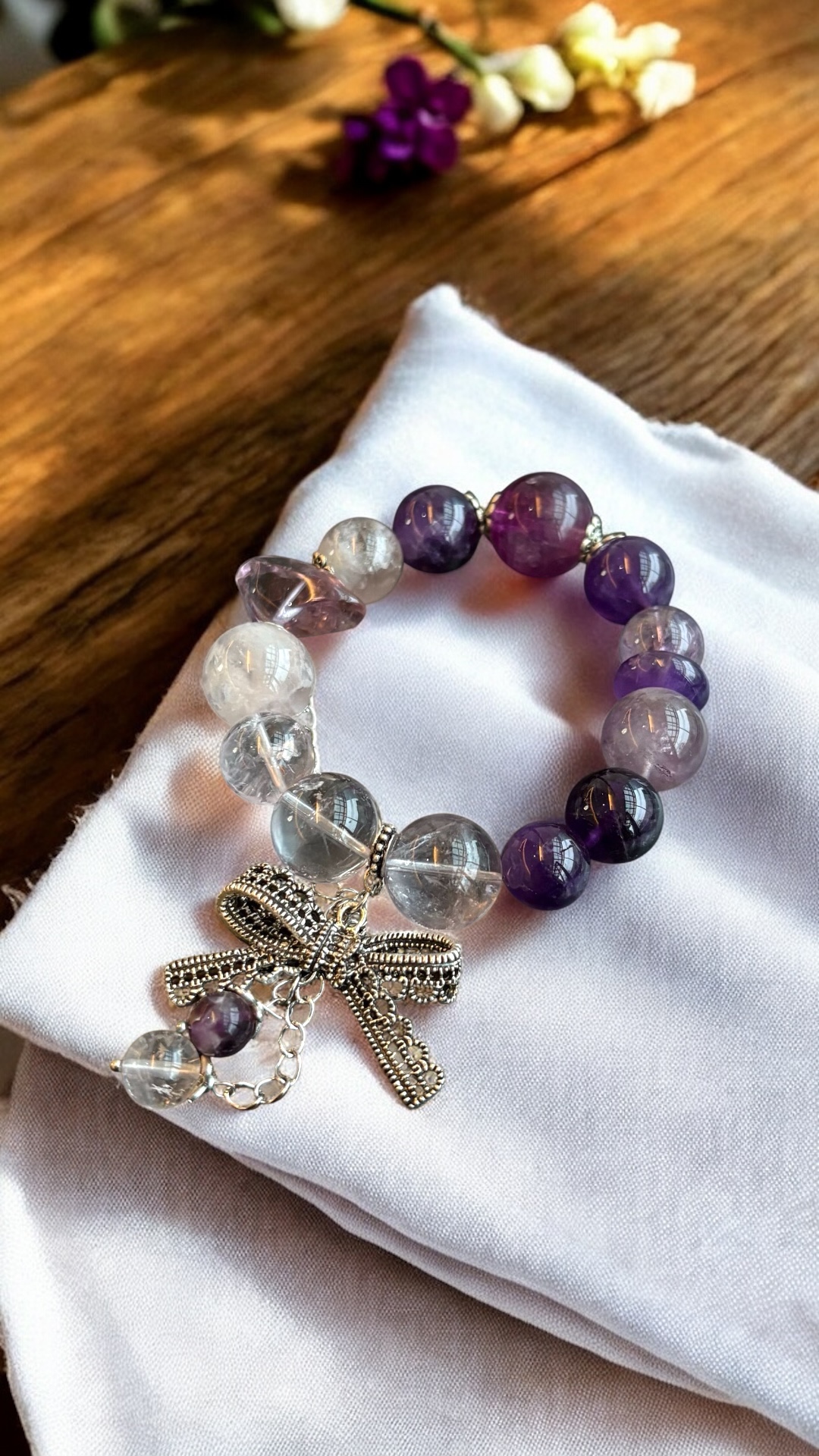 Amethyst and clear quartz gemstone bracelet featuring a silver bow charm and dangle chain on white fabric
