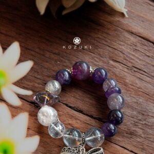 Amethyst and pink amethyst crystal bracelet with silver bow charm, styled on wooden background with soft floral light
