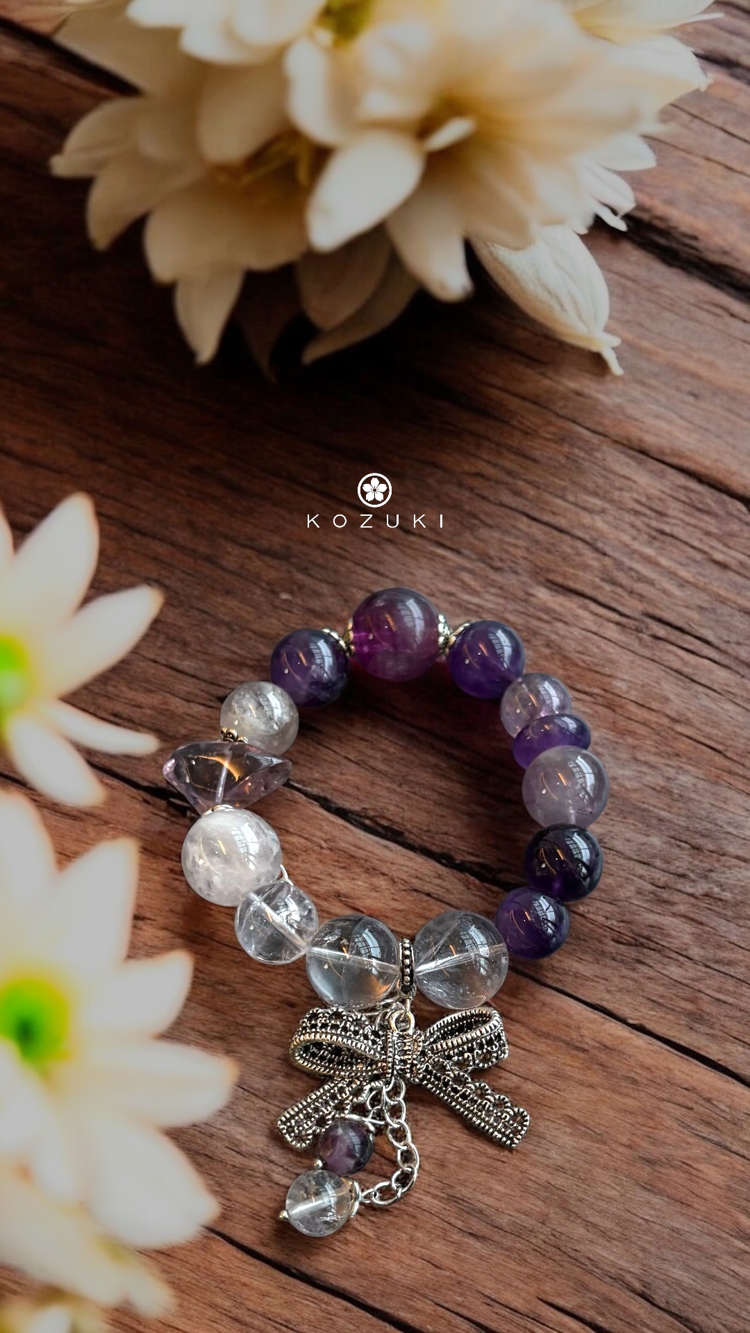 Amethyst and pink amethyst crystal bracelet with silver bow charm, styled on wooden background with soft floral light